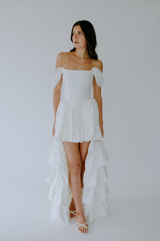 Woman wearing a white off-shoulder dress with ruffled layers on a plain background