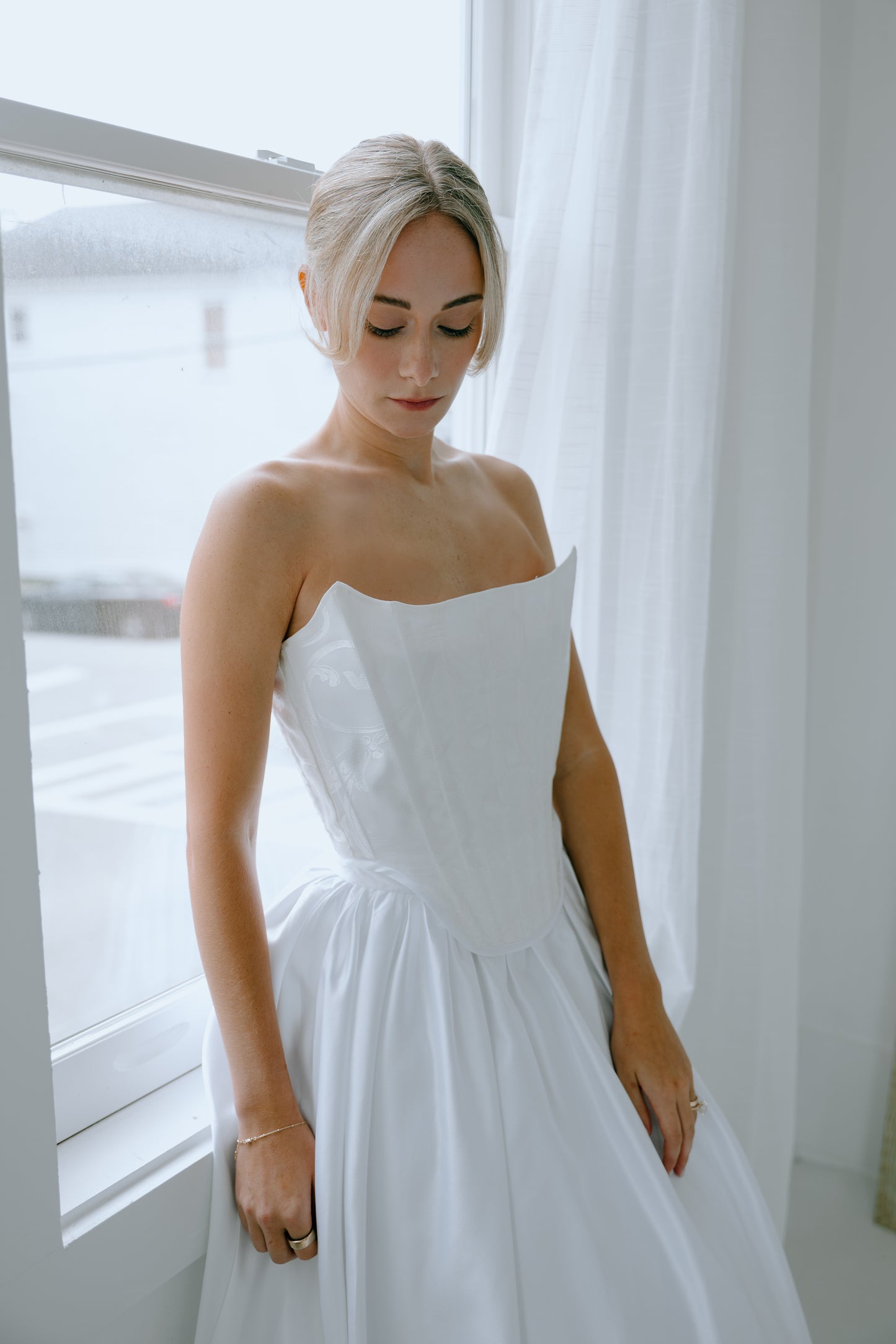 Woman in a white wedding dress standing by a window with white curtains.